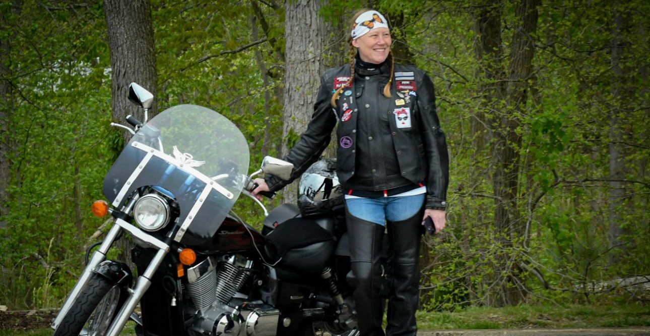 photo of a woman standing in the woods next to a motorcycle